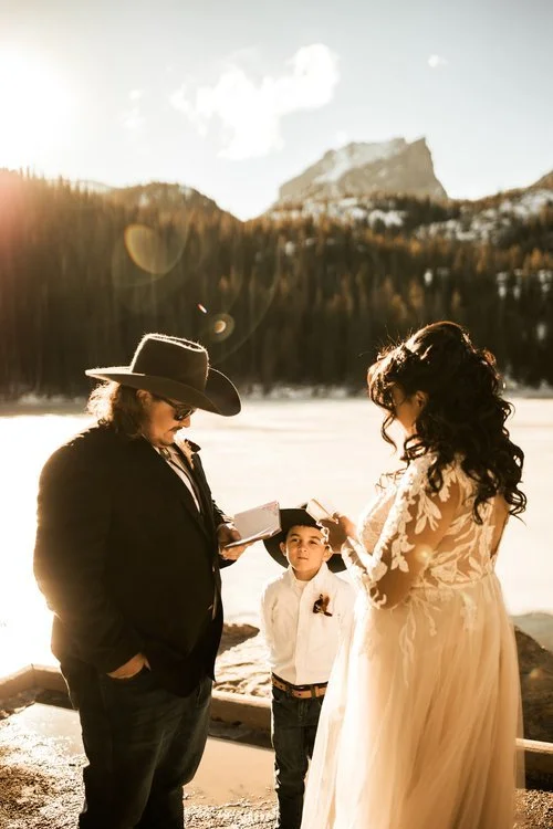 A couple reading their vows beside a frozen lake in Rocky Mountain National Park, a young boy in a suit watching between them as low sun breaks over the ridge